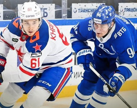 На изображении два хоккеиста в белой и синей форме на льду. Они активно борются за шайбу возле борта с рекламой. Игроки одеты в шлемы и защитную экипировку, их внимание сосредоточено на игре