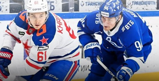 На изображении два хоккеиста в движении на льду. Игрок слева в форме с красными и синими элементами ведет шайбу, в то время как игрок справа в синей форме пытается блокировать его путь к воротам. На заднем плане рекламные баннеры