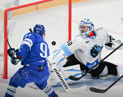 Хоккейный матч: игрок в синей форме атакует ворота, бросая шайбу, вратарь в бело-голубой экипировке старается отразить удар. Момент напряжённого противостояния на льду