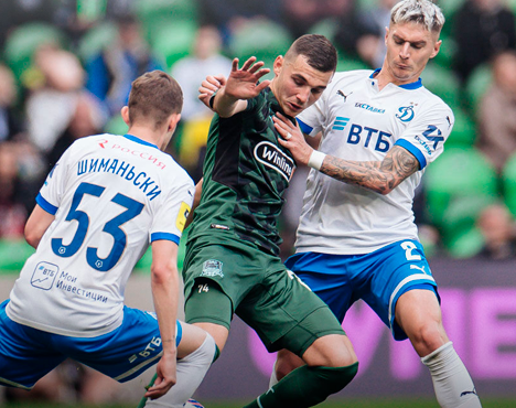 На изображении запечатлен момент футбольного матча: игрок в зеленой форме борется за мяч с двумя соперниками в бело-синей форме. Интенсивность игры видна по их действиям и эмоциям