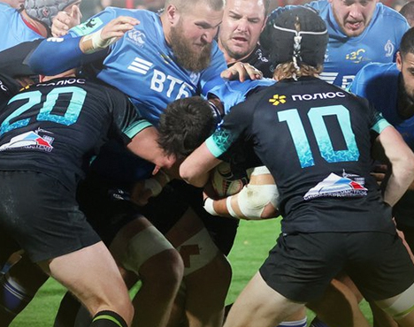 На изображении игроки двух команд сосредоточены в борьбе за мяч во время регбийного матча. Яркая и динамичная сцена подчеркивает напряженность и командный дух игры